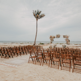 An outdoor beach wedding ceremony setup with wooden chairs and floral arch in Riviera Maya